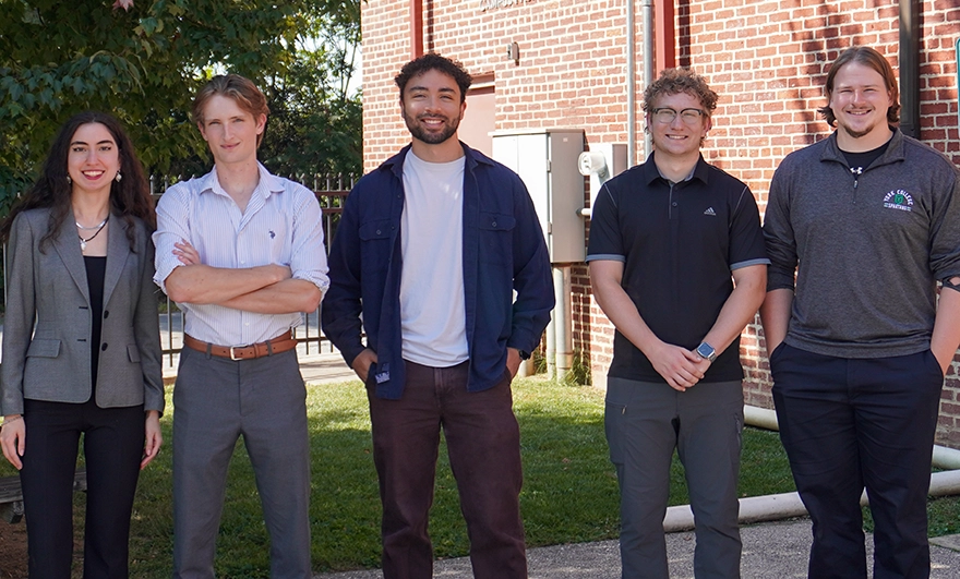 Five students, four men and one woman, standing outside in front of a brick building