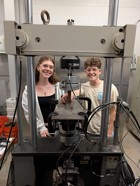 Two students wearing safety glasses smile beside a large hydraulic testing press in a lab.