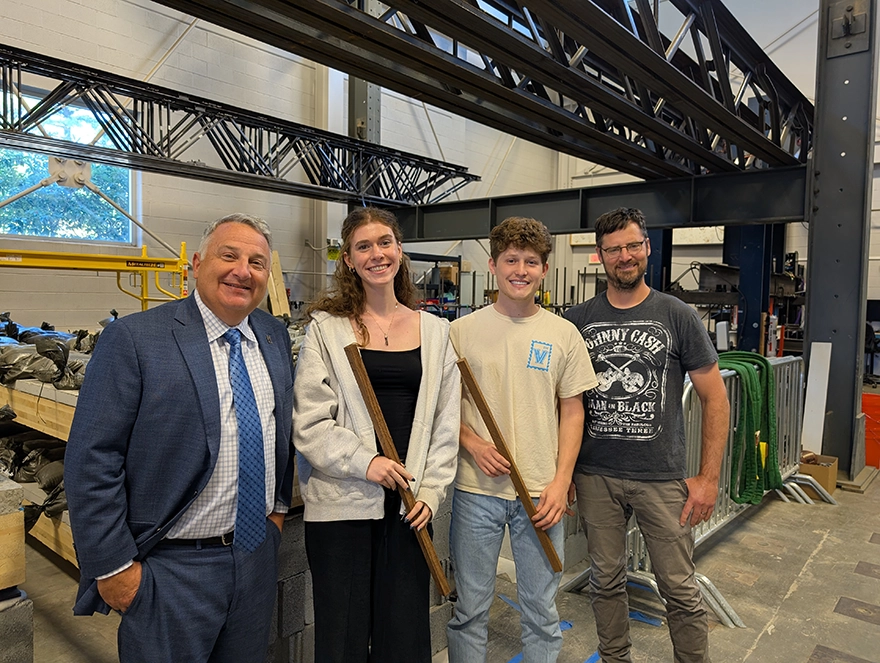 Four people pose in an industrial workshop beneath large steel trusses, with two holding long wooden strips.